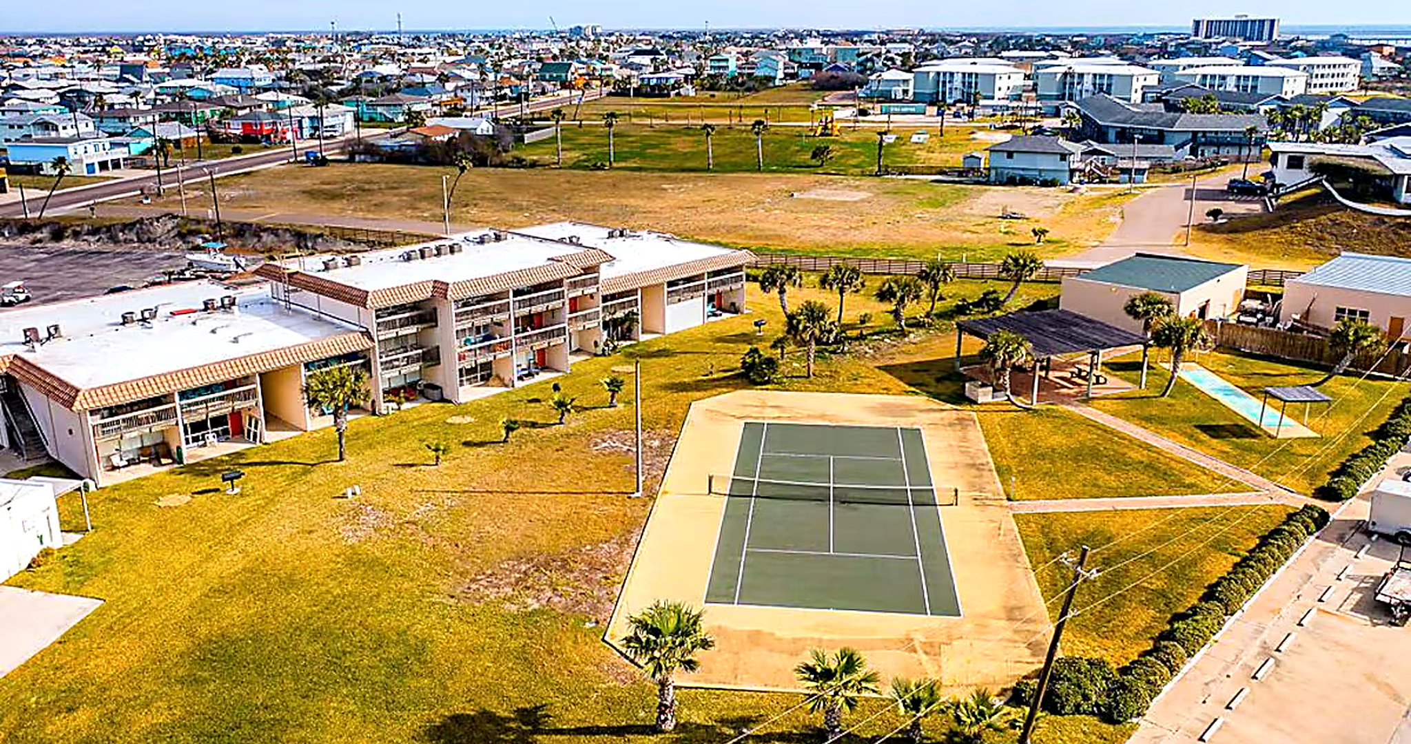 Port Aransas condo complex with pool, tennis court, lawns, and palm trees near the beach
